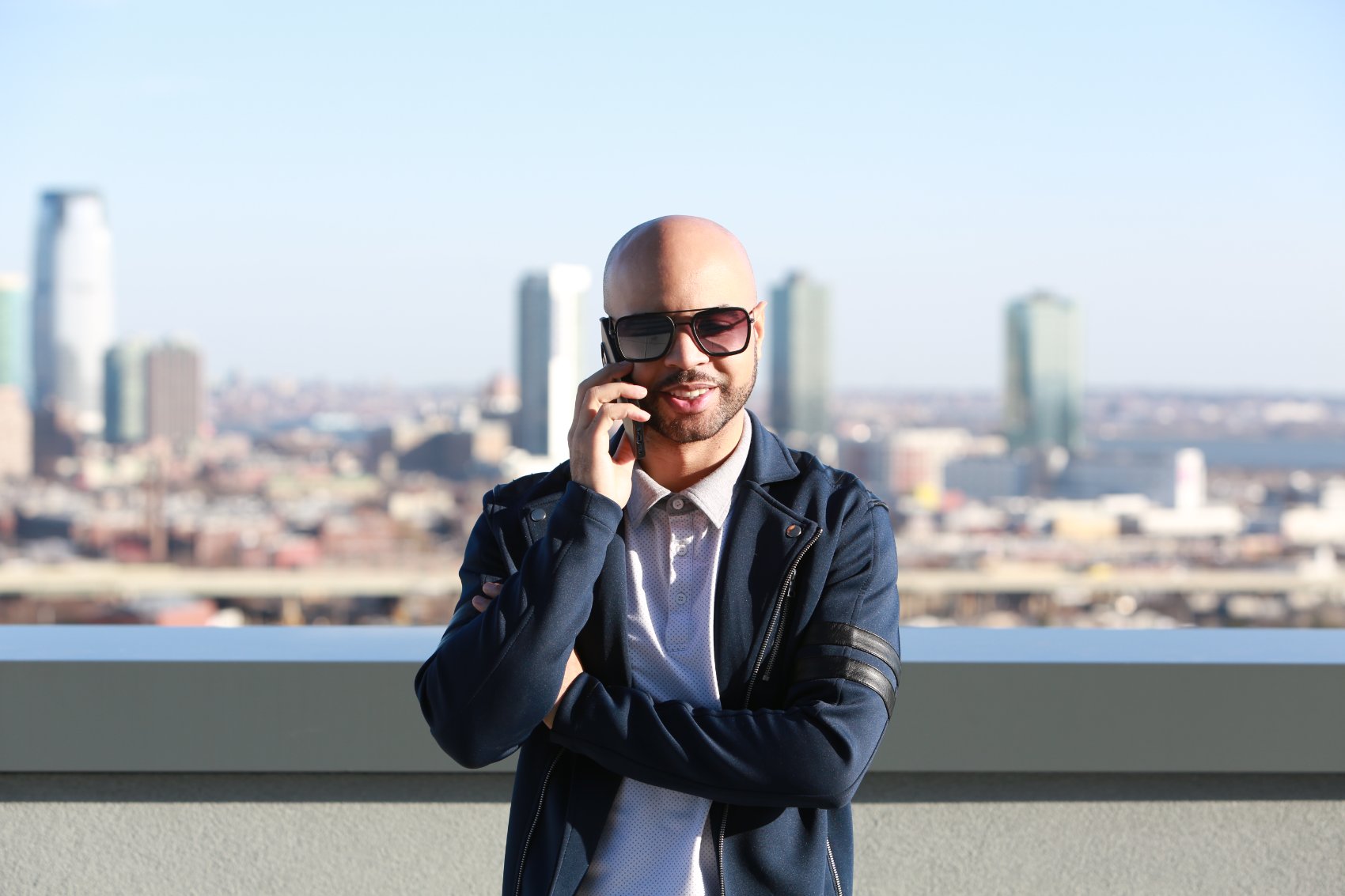 Cristo Hernandez, founder and CEO of ARC Property Management, smiling while on a phone call on a rooftop with the Jersey City skyline behind him.